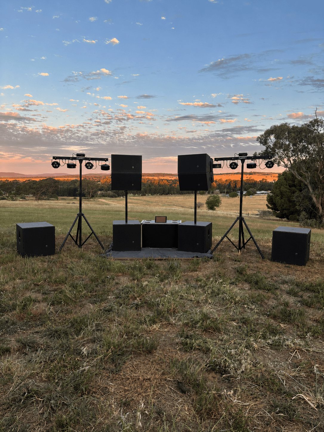 LumixAV full PA and lighting rig set up in a paddock at sunset — two tops, two subs, GigBAR lighting and a DJ booth